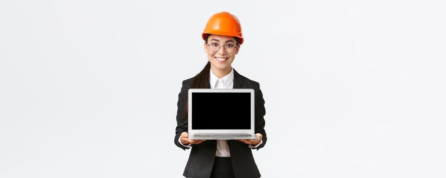 Smiling professional female asian engineer introduce construction plan to investors or clients, standing in safety helmet and suit showing laptop screen with pleased smile, white background.