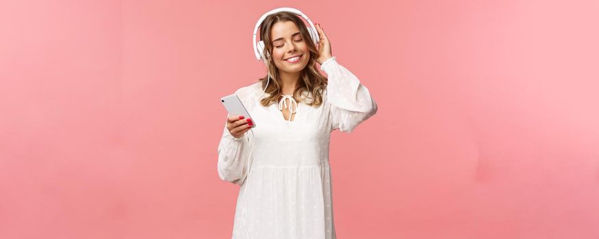 Portrait of tender happy and cute blond girl in white dress, close eyes and smiling pleased as listening music in headphones, feeling warmth and happiness enjoying favorite track, hold mobile phone.