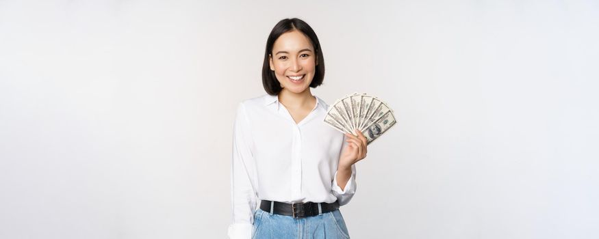 Credit and loan concept. Smiling young asian woman holding cash dollars and looking happy at camera, white background.