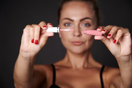 Focus on a moisturizing pink liquid lip balm gloss in the hands of a blurred beautiful Caucasian blonde woman isolated over black background with copy ad space.