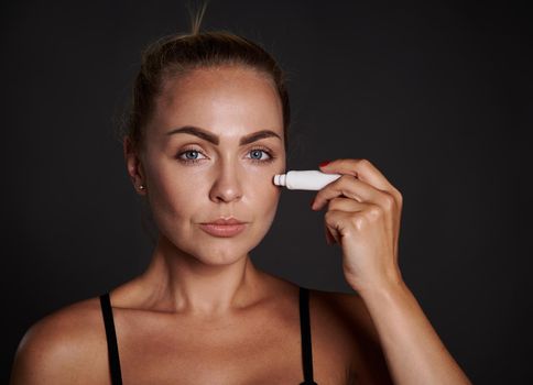 Beautiful middle aged woman applying anti puffiness anti wrinkles moisturizer under eyes. Youth concept and prevention of first signs of aging process.