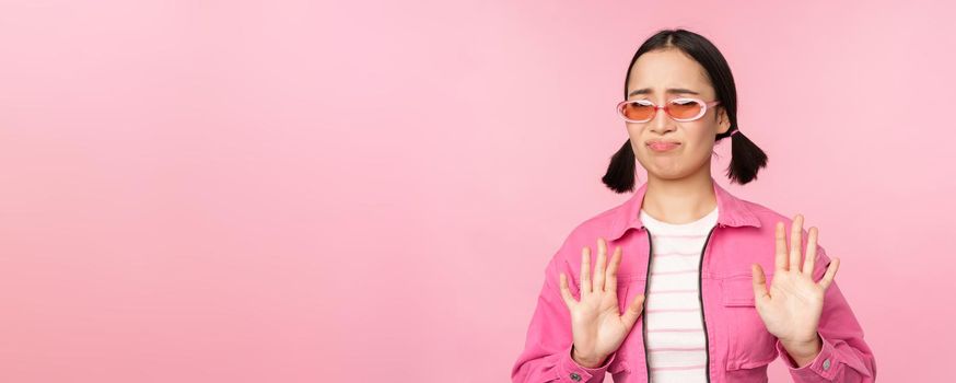 No thanks. Disgusted asian girl in sunglasses, rejecting smth, shaking hands to refuse, decline, standing over pink background.