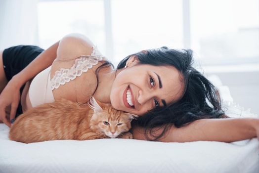 Close up view. Attractive blonde resting on the white bed with her cute kitten.