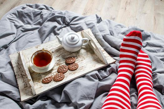 Fragment of legs in red striped socks. interior and home coziness concept. Top view. A cup of tea, a teapot with herbal tea, sugar bowl on a wooden white tray on the bed. Porcelain cup