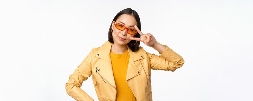 Stylish asian girl in sunglasses, looks cool and trendy, shows peace, v-sign gesture, stands over white background. Copy space