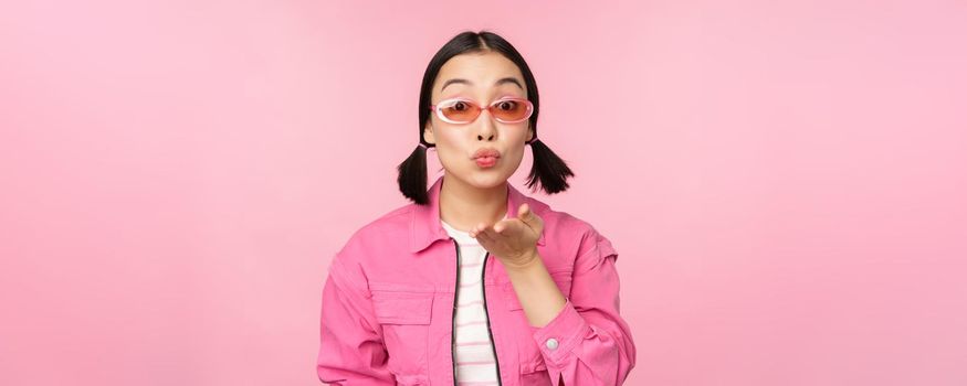 Stylish k-pop girl in sunglasses, asian woman blowing air kiss at camera, pucker lips, mwah gesture, standing over pink studio background. Copy space