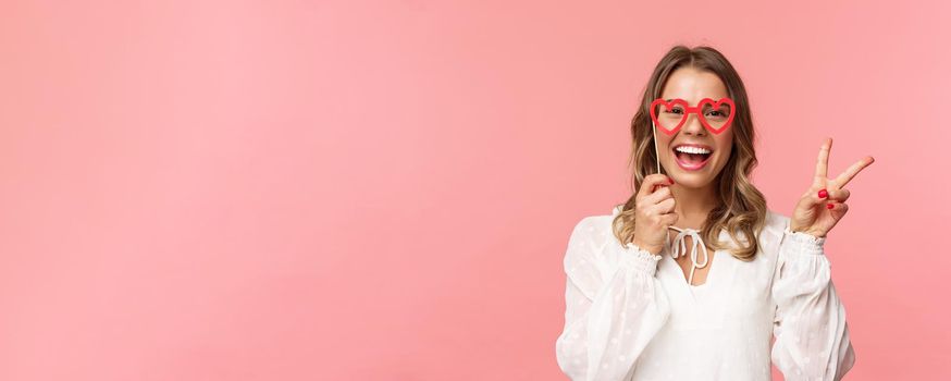 Spring, happiness and celebration concept. Close-up portrait of upbeat blond girl at party, wear white dress holding heart-shaped glasses mask over eyes and make peace sign cheerful.