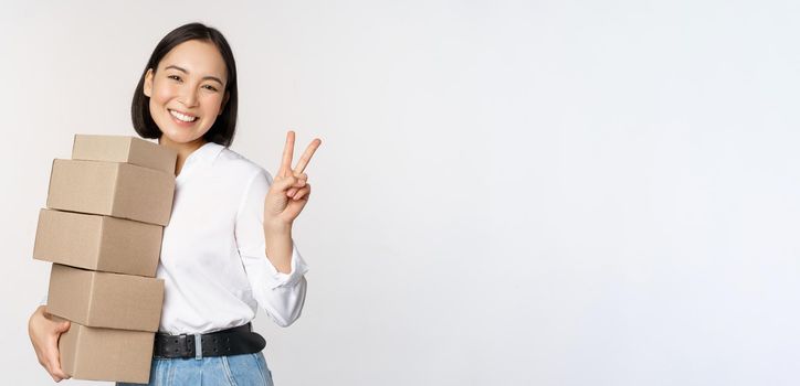 Smiling korean woman with boxes showing v-sign, peace gesture, standing over white background.