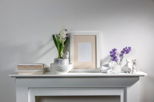 White hyacinth in a large porcelain bowl, books, figurines of hares and a bird, an empty photo frame are on the fireplace against the white wall. Layout.