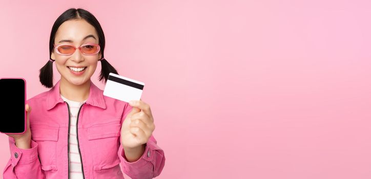 Image of smiling korean woman showing credit card and mobile phone screen, smartphone application interface, paying online, shopping contactless, standing over pink background.