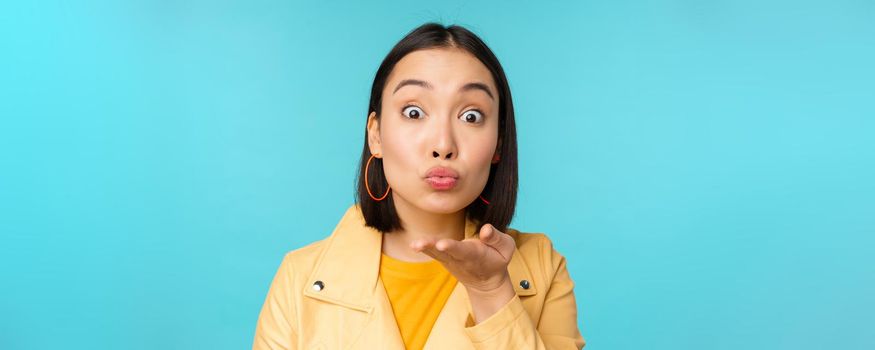 Close up portrait of funny asian girl sending air kiss, blowing at camera with popped eyes, standing over blue background.