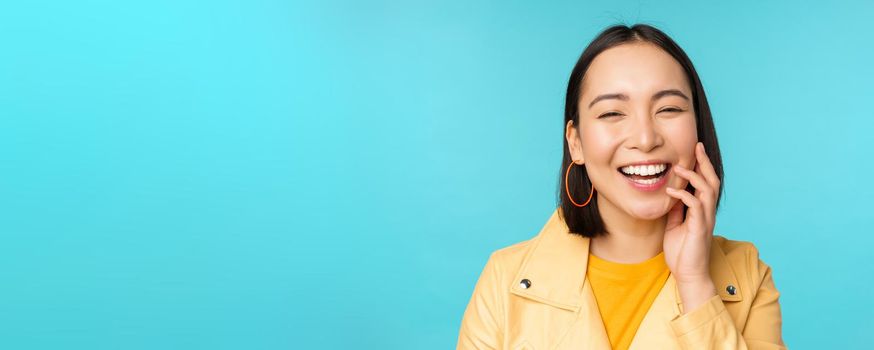 Close up portrait of natural asian girl laughing, smiling and looking happy, standing over blue background. Copy space