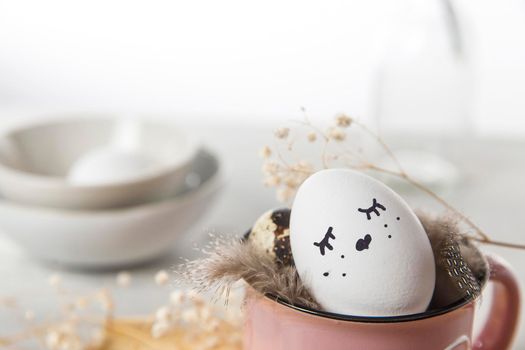 Easter concept. A white egg with copper wire ears and a painted rabbit sleeping face in a cup. Partridge eggs and feathers.