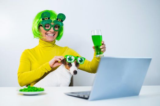 A young woman in a green wig and cheerful glasses drinks beer and bites glazed nuts. A girl sits with a dog at a table and celebrates st patrick's day online chatting with friends on a laptop