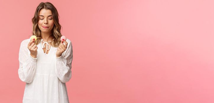 Holidays, spring and party concept. Portrait of cute romantic blond girl in white dress, licking lips as tempting to eat delicious dessert, holding two macarons and look pleased, pink background.