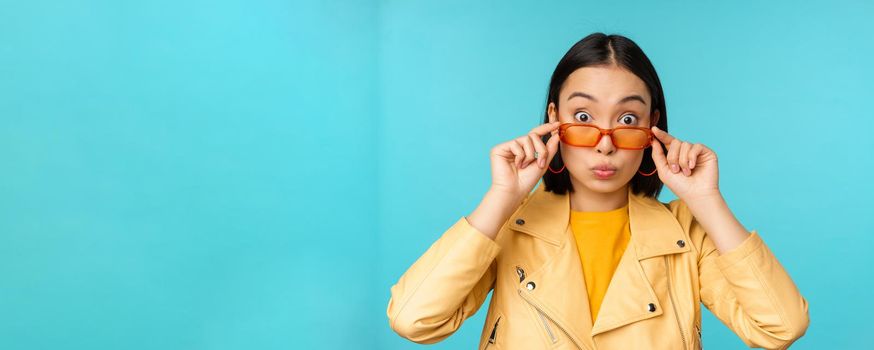 Close up portrait of asian girl in sunglasses, looking happy and surprised, excitement on face, standing over blue background.