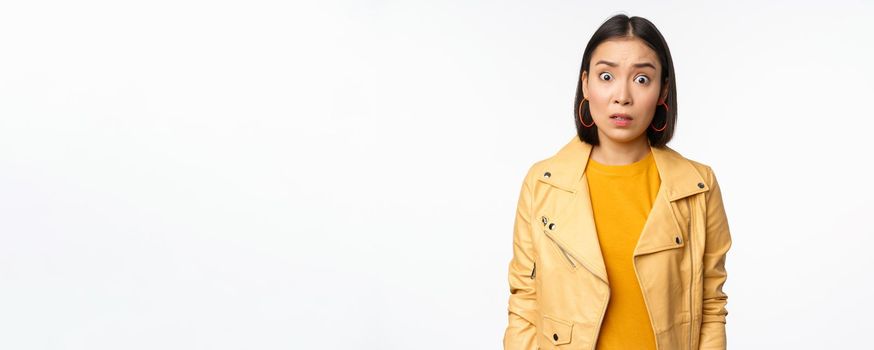 Image of shocked, confused asian woman, staring frustrated and speechless at camera, raising eyebrows and looking concerned, standing over white background.