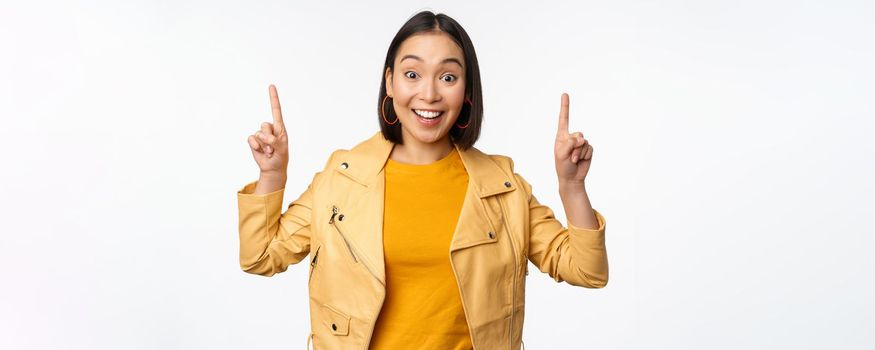 Enthusiastic asian girl pointing fingers up, showing advertisement on top, smiling happy, demonstrating promo offer or banner, standing over white background.