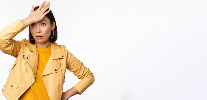 Annoyed and tired asian girl, slap forehead, facepalm and eyeroll with bothered face expression, standing in casual clothes over white studio background.