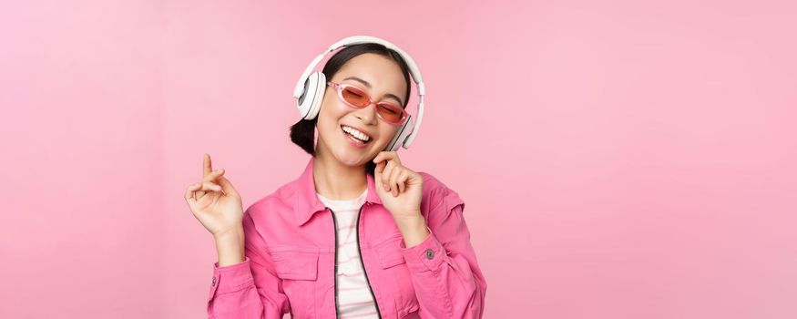 Dancing stylish asian girl listening music in headphones, posing against pink background.