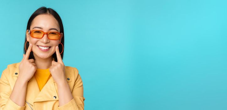 Close up portrait of asian young woman in sunglasses, smiling and looking romantic, standing happy over blue background. Copy space