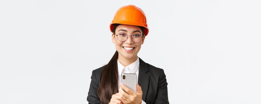 Close-up of successful female chief engineer, construction architect in safety helmet and business suit, glasses, looking at camera satisfied, smiling pleased as using mobile phone.