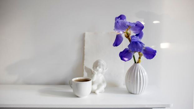 Blue iris in a corrugated vase with a piece of craft paper on a white table. Figurine of the angel. Cup of tea. Place for text