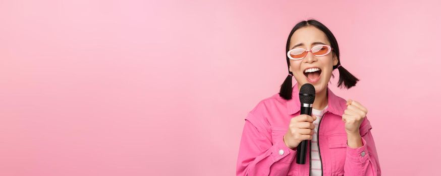 Happy beautiful asian girl singing with mic, using microphone, enjoying karaoke, posing against pink studio background.