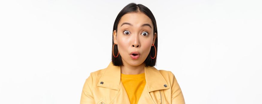 Close up of impressed asian girl, say wow, looking amazed at camera, standing in casual clothes over white background. Copy space
