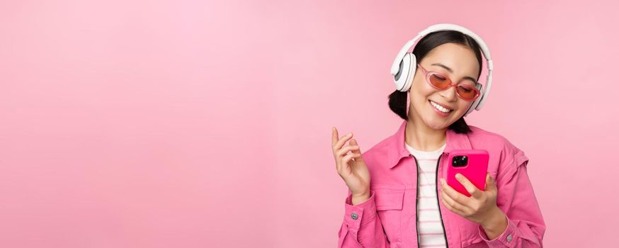 Stylish asian girl dancing with smartphone, listening music in headphones on mobile phone app, smiling and laughing, posing against pink background.