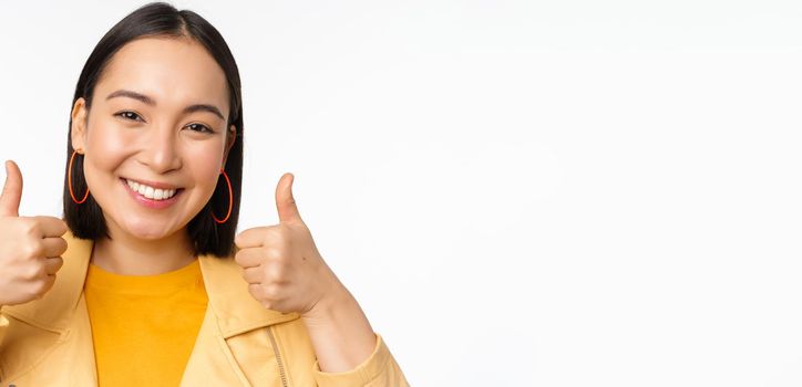 Smiling asian girl showing thumbs up, looking pleased, approve smth, standing over white background. Copy space