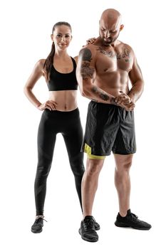 Strong bald, tattooed fellow in black shorts and sneakers with charming brunette lady in leggings and top are posing isolated on white background and looking at the camera. Fitness couple, chic muscular bodies, gym concept. The love story.