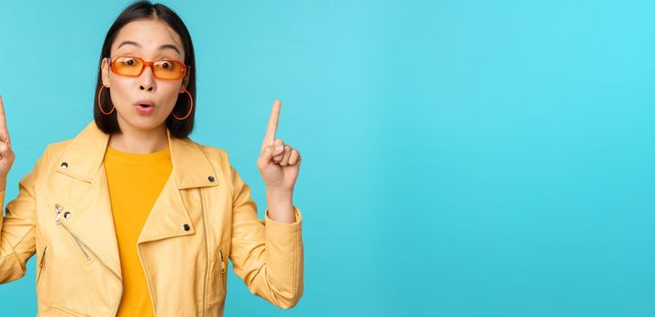 Enthusiastic asian girl in sunglasses, points fingers up, shows banner or logo on top, stands over blue background. Copy space