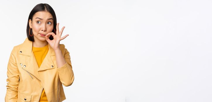 Image of young asian woman seal lips, silence gesture, zipping her mouth with promise, standing over white background.