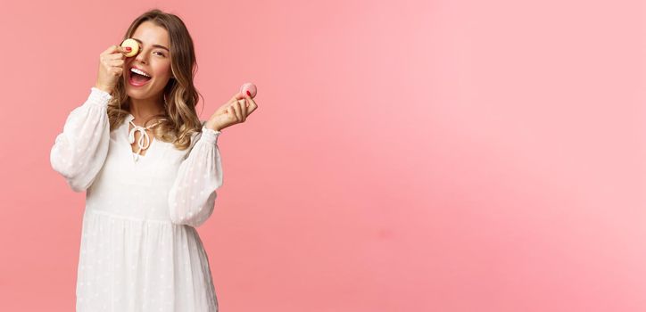 Holidays, spring and party concept. Portrait of carefree, tender and feminine girl in white dress, holding macarons dessert over eye, feeling cheerful, eating sweets, tasty desserts, pink background.