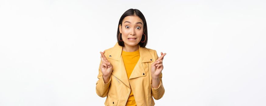 Portrait of excited asian woman looks hopeful, wishing, praying or begging, waiting for news, standing over white background, smiling enthusiastic.