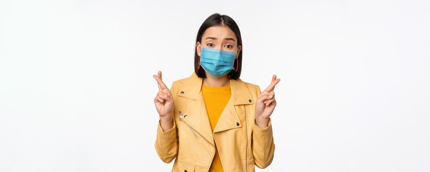 Hopeful asian girl in medical face mask, cross fingers, making wish, hoping, praying for smth, standing with anticipation over white studio background.