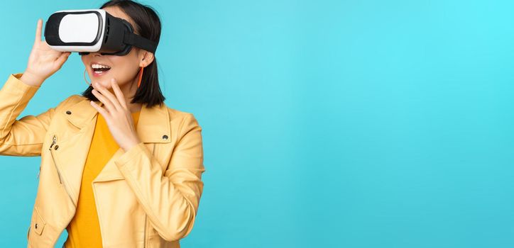 Portrait of happy asian female model using VR headset, smiling and laughing in virtual reality glasses, standing over blue background.