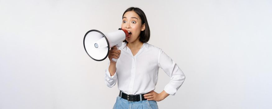 Image of young woman, korean activist, recruiter screaming in megaphone, searching, shouting at loudspeaker, standing over white background.