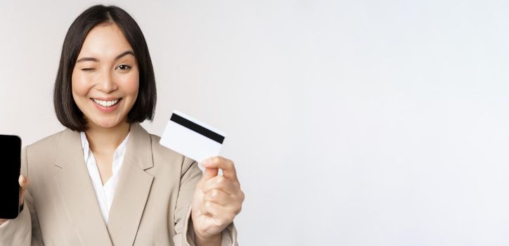 Smiling corporate woman in suit, showing mobile phone screen and app on mobile phone, smartphone screen, standing over white background.