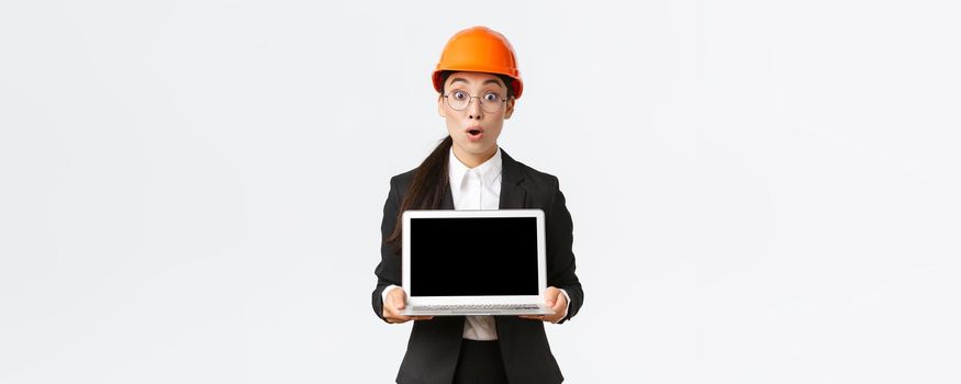 Impressed and excited asian female engineer showing amazing results on graph, industrial technician in safety helmet and suit showing laptop screen with amazed expression, white background.