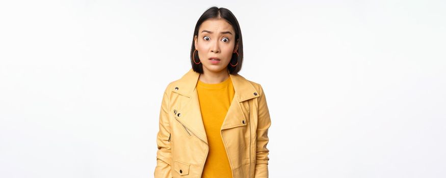 Image of shocked, confused asian woman, staring frustrated and speechless at camera, raising eyebrows and looking concerned, standing over white background.