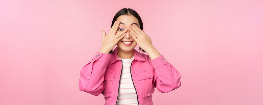 Portrait of asian girl peeks with excitement through fingers, covers eyes, seeing surprise, standing over pink background. Copy space