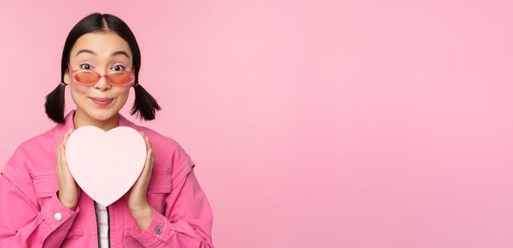 Beautiful asian girl smiling happy, showing heart gift box and looking excited at camera, standing over pink romantic background.