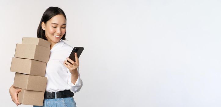 Image of young asian woman holding boxes, customer orders and looking at mobile phone, standing over white background.