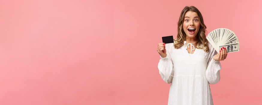 Portrait of excited happy good-looking blond girl in white dress, winning money, placed good bet, made deal, holding dollars money and credit card, smiling amused at camera, pink background.
