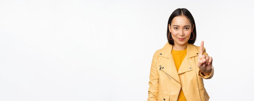 Rule number one. Confident smiling asian woman showing index finger, stop, prohibit gesture, disapprove smth, standing over white background.