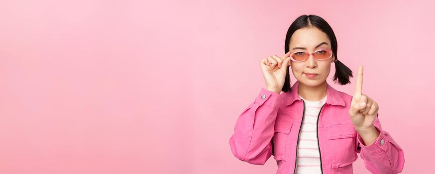Image of asian girl with serious face, showing prohibit, scolding gesture, shaking finger pointing up, express disapproval, standing over pink background.