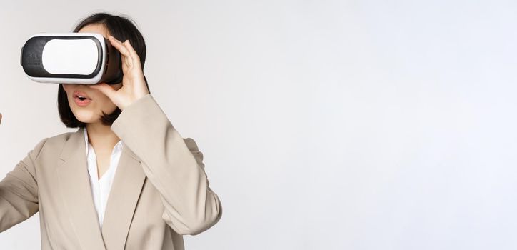 Amazed business woman in suit using virtual reality glasses, looking amazed in vr headset, standing over white background.