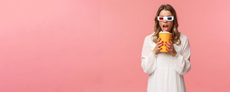 Leisure, going-out and spring concept. Portrait of cute blond funny girl in cinema, eating popcorn from box without hands, looking aside suspicious, wearing 3d glasses, pink background.